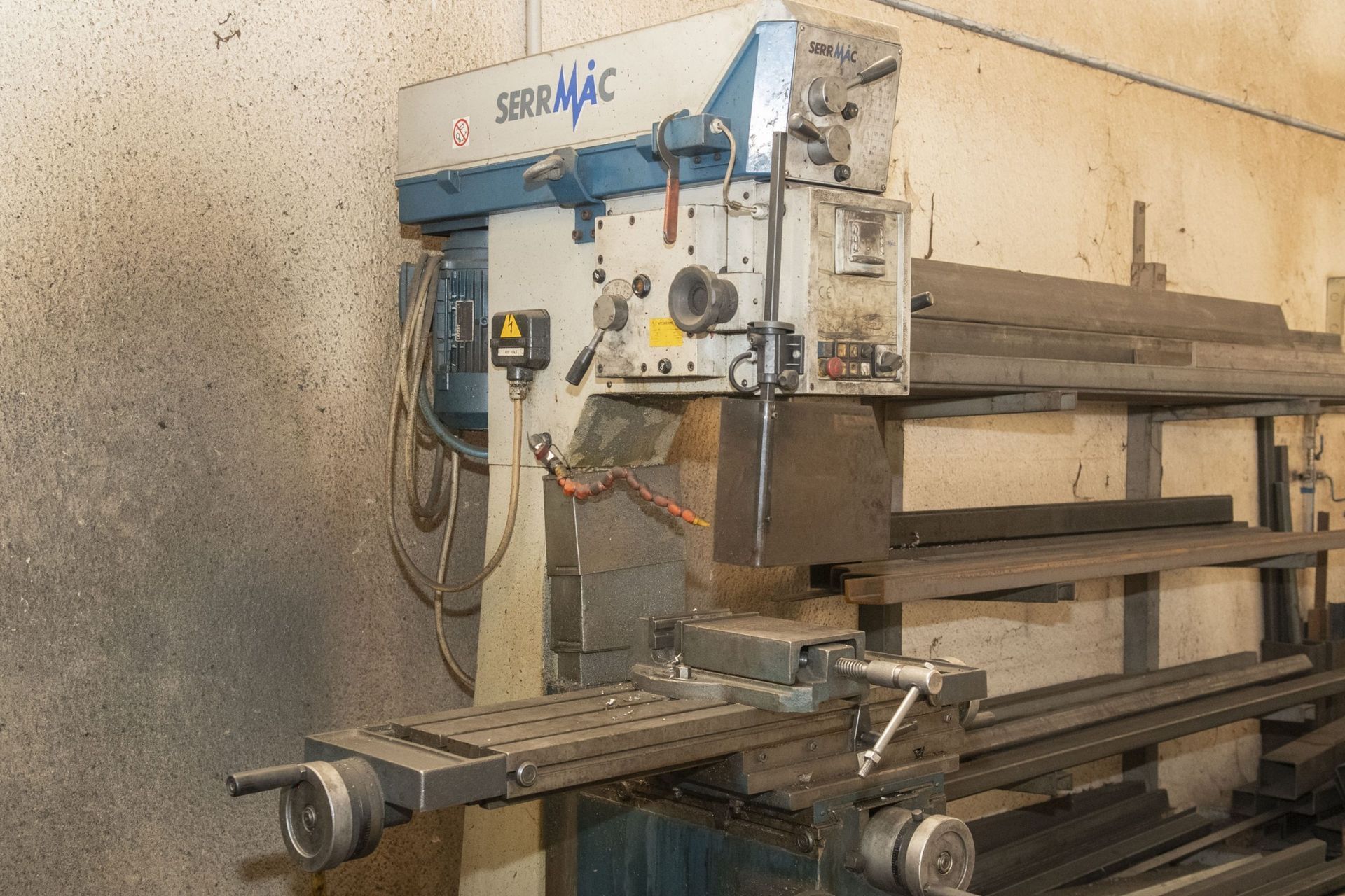 Una fresatrice industriale Serrmac di colore grigio e blu è appoggiata a una parete con superficie ruvida, accanto a una scaffalatura metallica con materiali in magazzino.