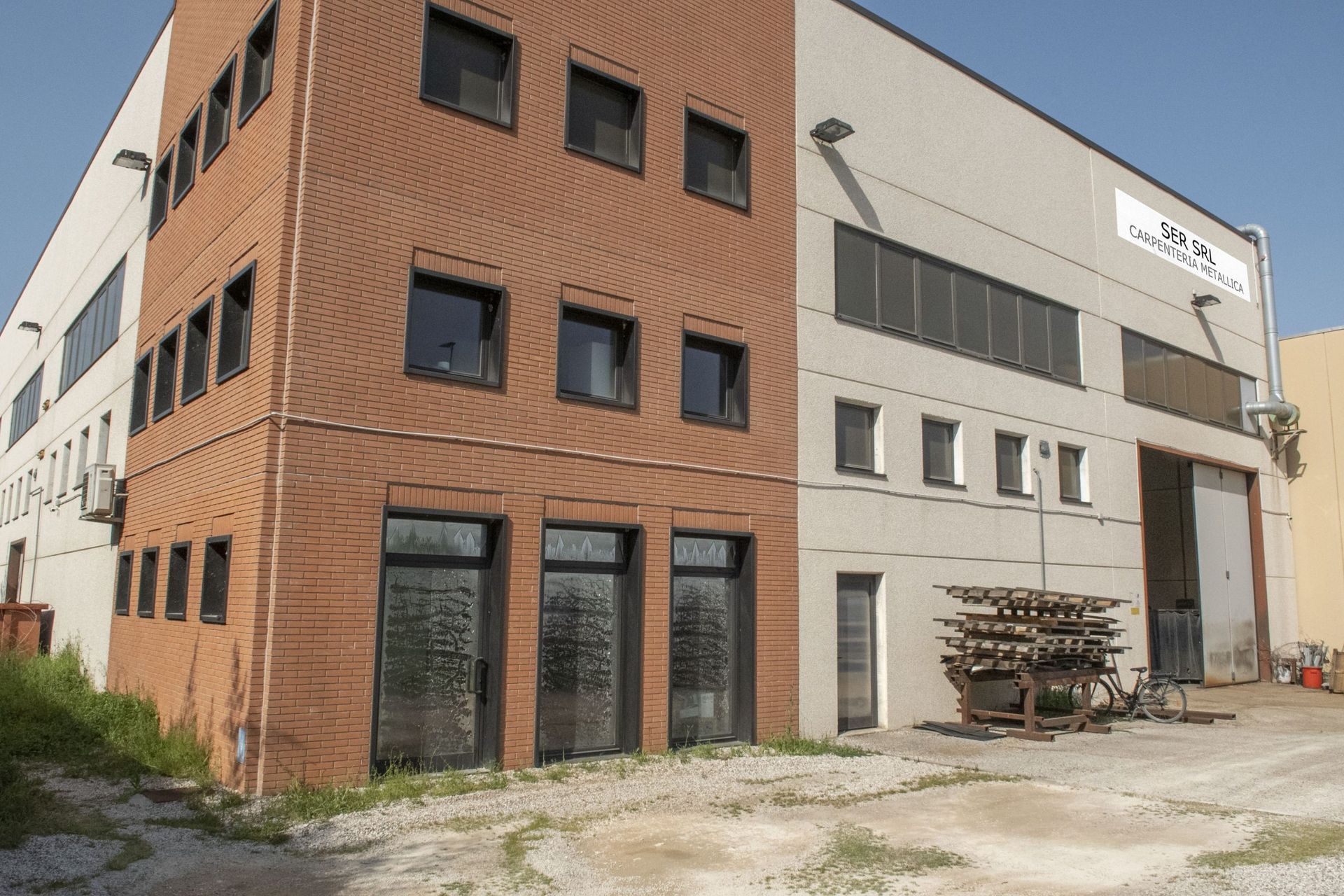 Un edificio a due piani con facciata in mattoni di terracotta a sinistra e pannelli industriali grigio chiaro a destra.