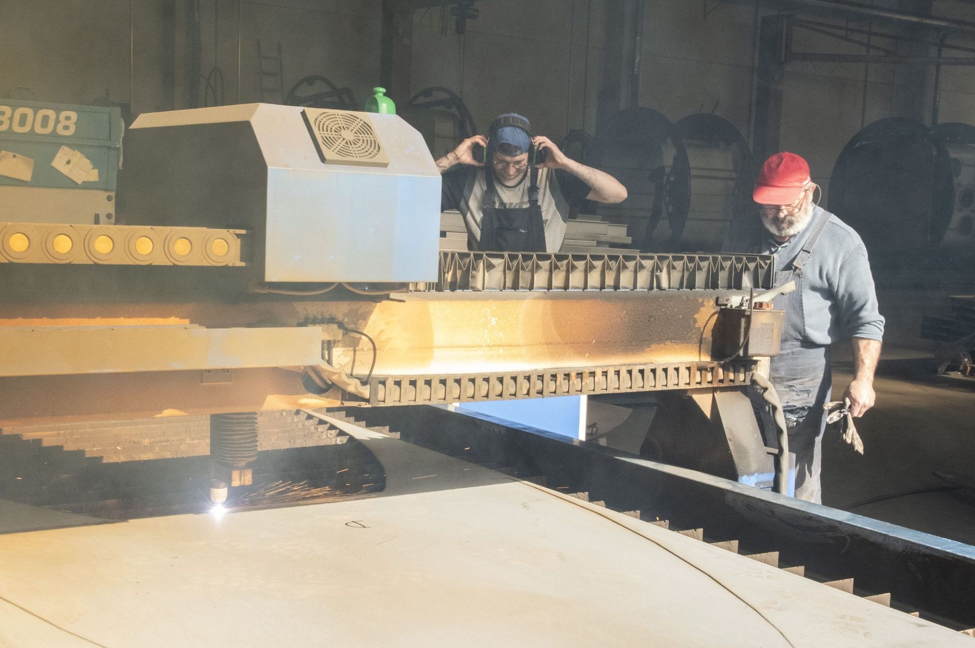 In un'officina industriale, due operai monitorano una grande macchina a controllo numerico (CNC) che taglia il metallo, illuminata da una scintilla.