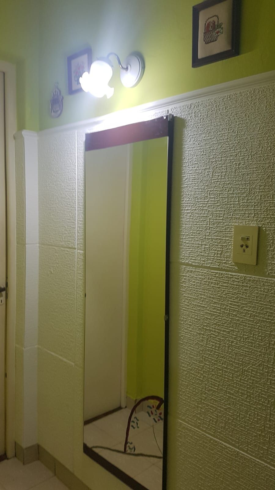 Espejo sobre pared texturizada, iluminado por una lámpara de dos focos. Paredes verdes, puerta y un cuadro enmarcado.