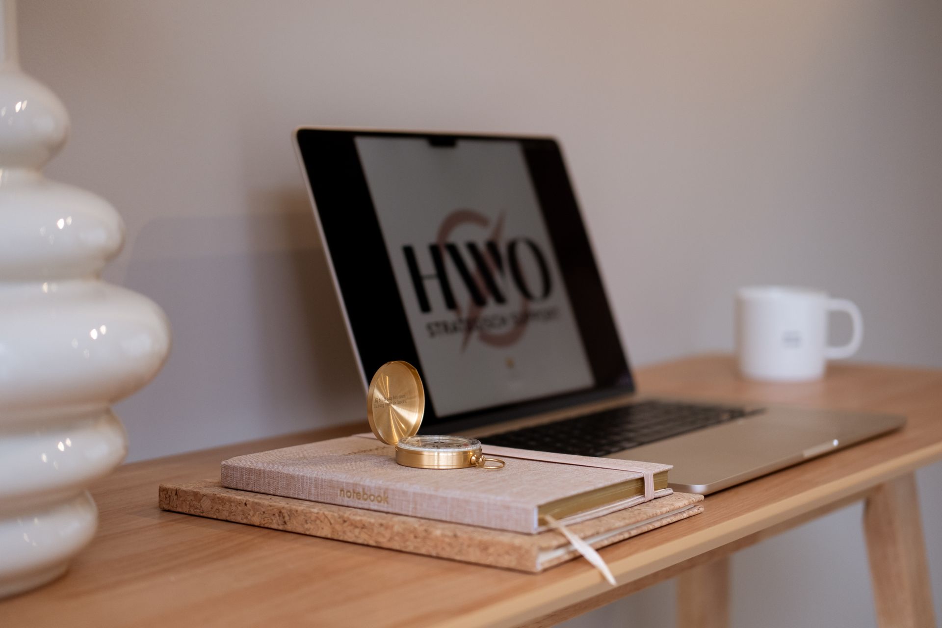 Laptop, notitieboek, kompas en mok op een houten bureau met een witte lamp, in een lichtgekleurde kamer.