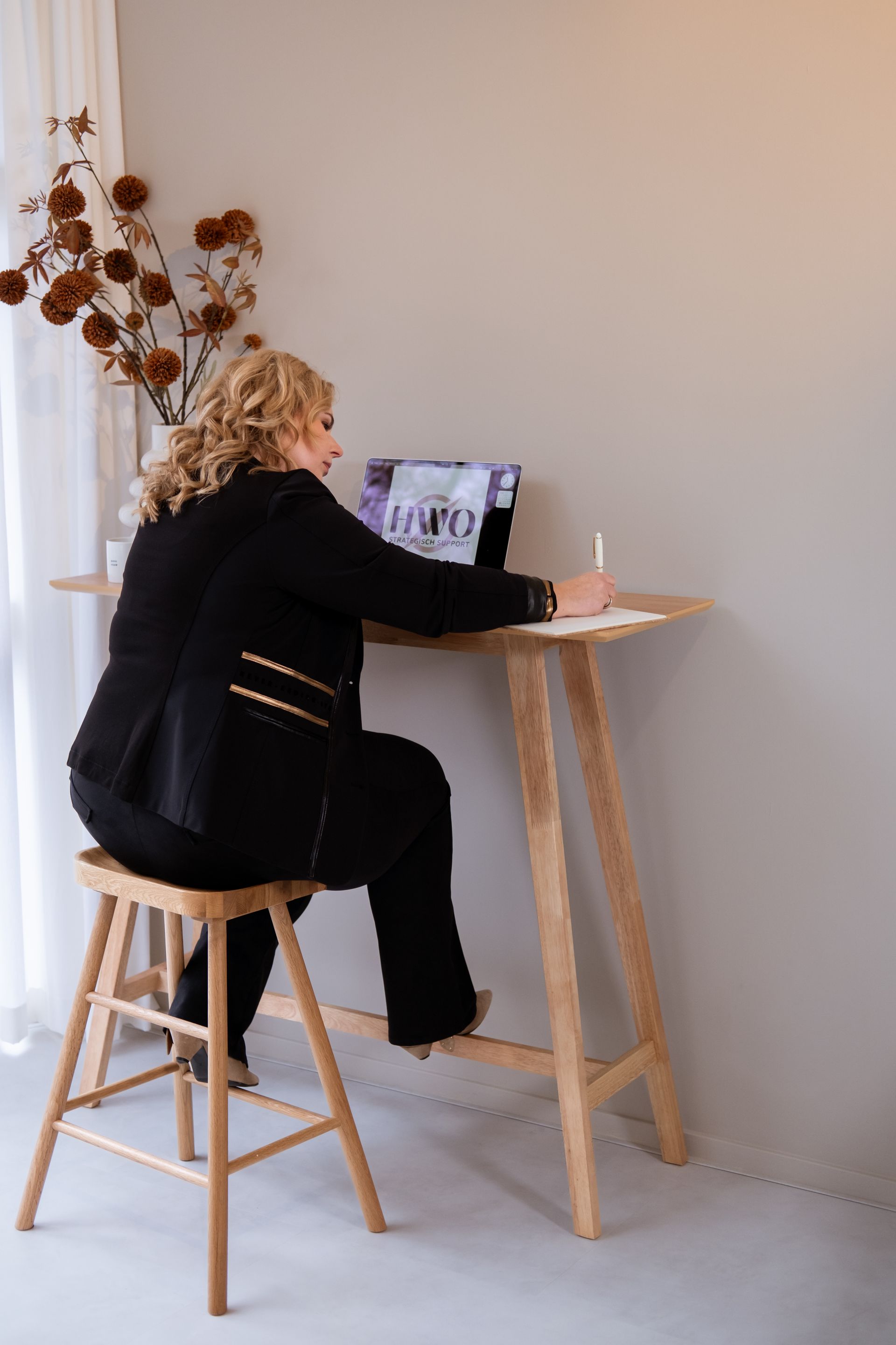Vrouw in zwart pak aan een hoog houten bureau, schrijvend op een tablet. Beige muur, gedroogde bloemen in een vaas.