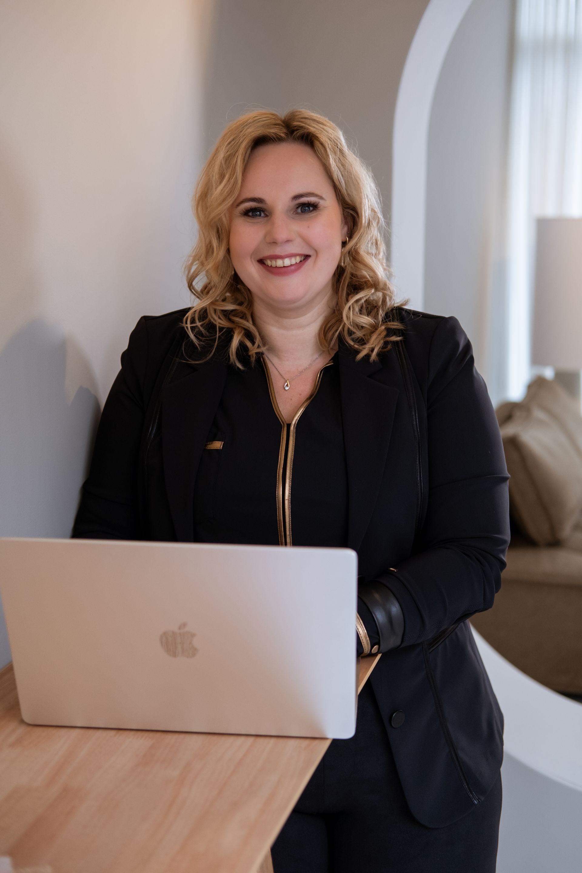 Een vrouw in een zwart pak glimlacht terwijl ze met haar laptop tegen een tafel leunt, binnen.