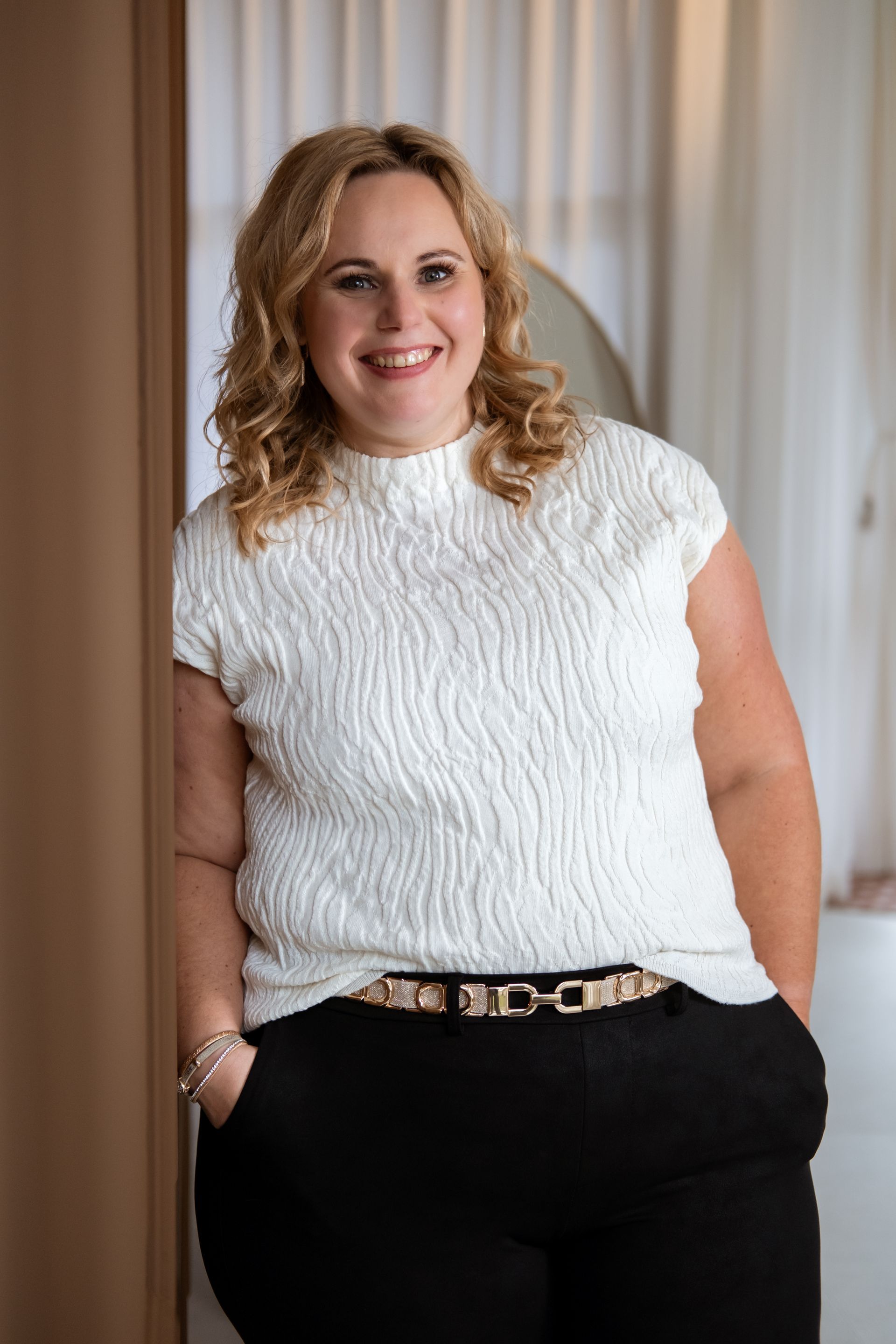 Vrouw in een witte top met textuur en een zwarte broek, lachend met haar handen in haar zakken.