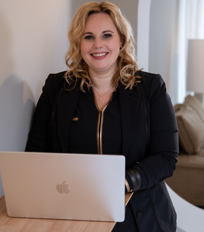 Een glimlachende vrouw, leunend tegen de muur, typt op een laptop en draagt een zwarte blazer.
