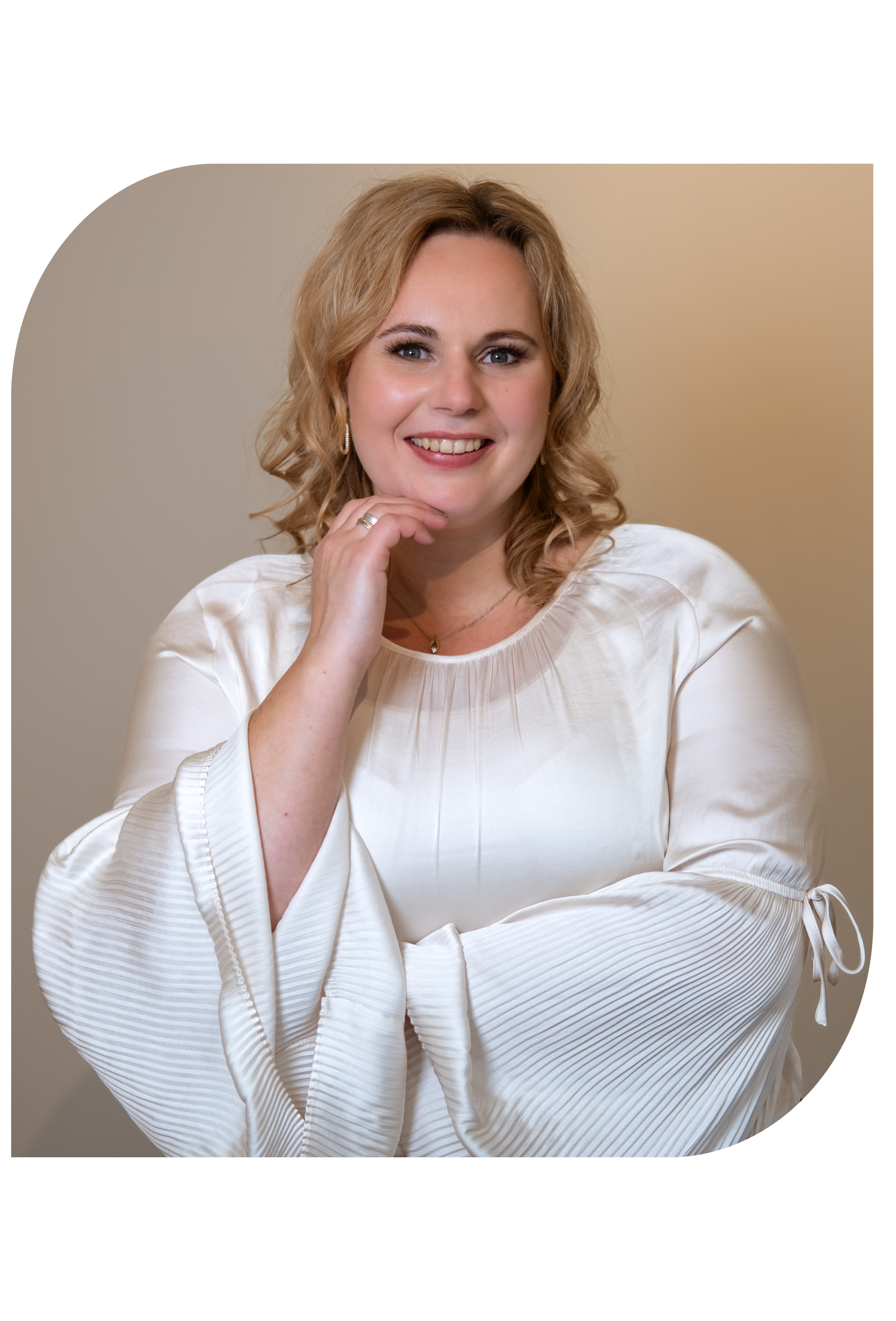 Vrouw in witte top, lachend, hand aan haar kin, binnen, neutrale achtergrond.