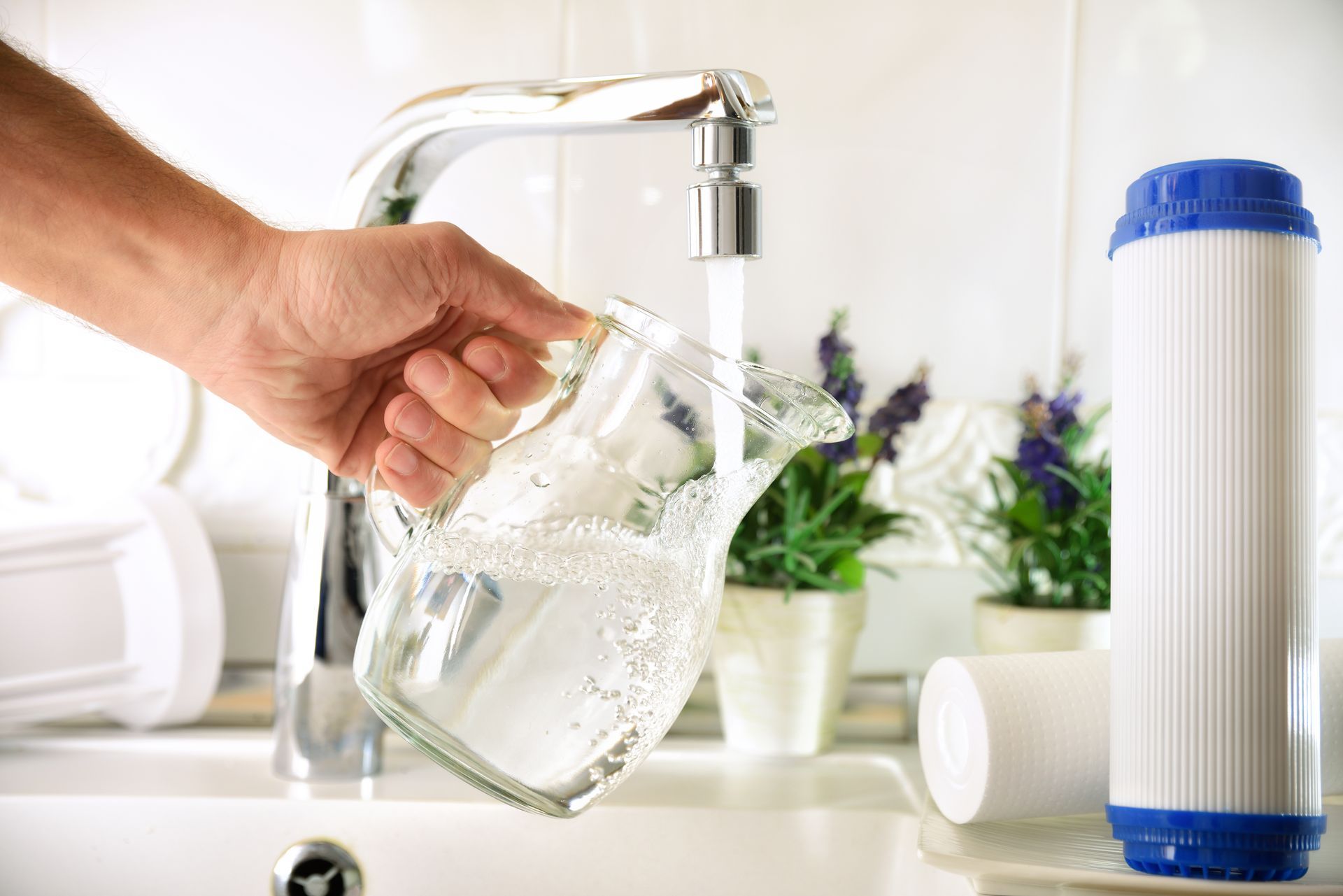 Hand filling a glass pitcher with water from a kitchen faucet beside water filter cartridges.