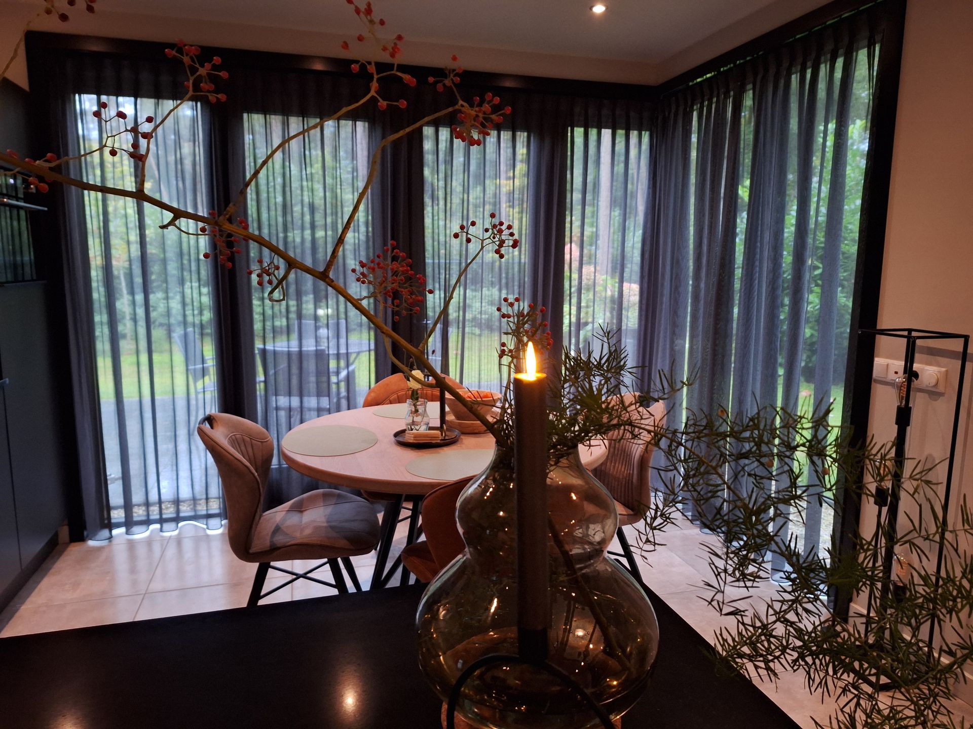 Eetgedeelte met transparante gordijnen, een kaarsverlicht middelpunt en uitzicht op groen.