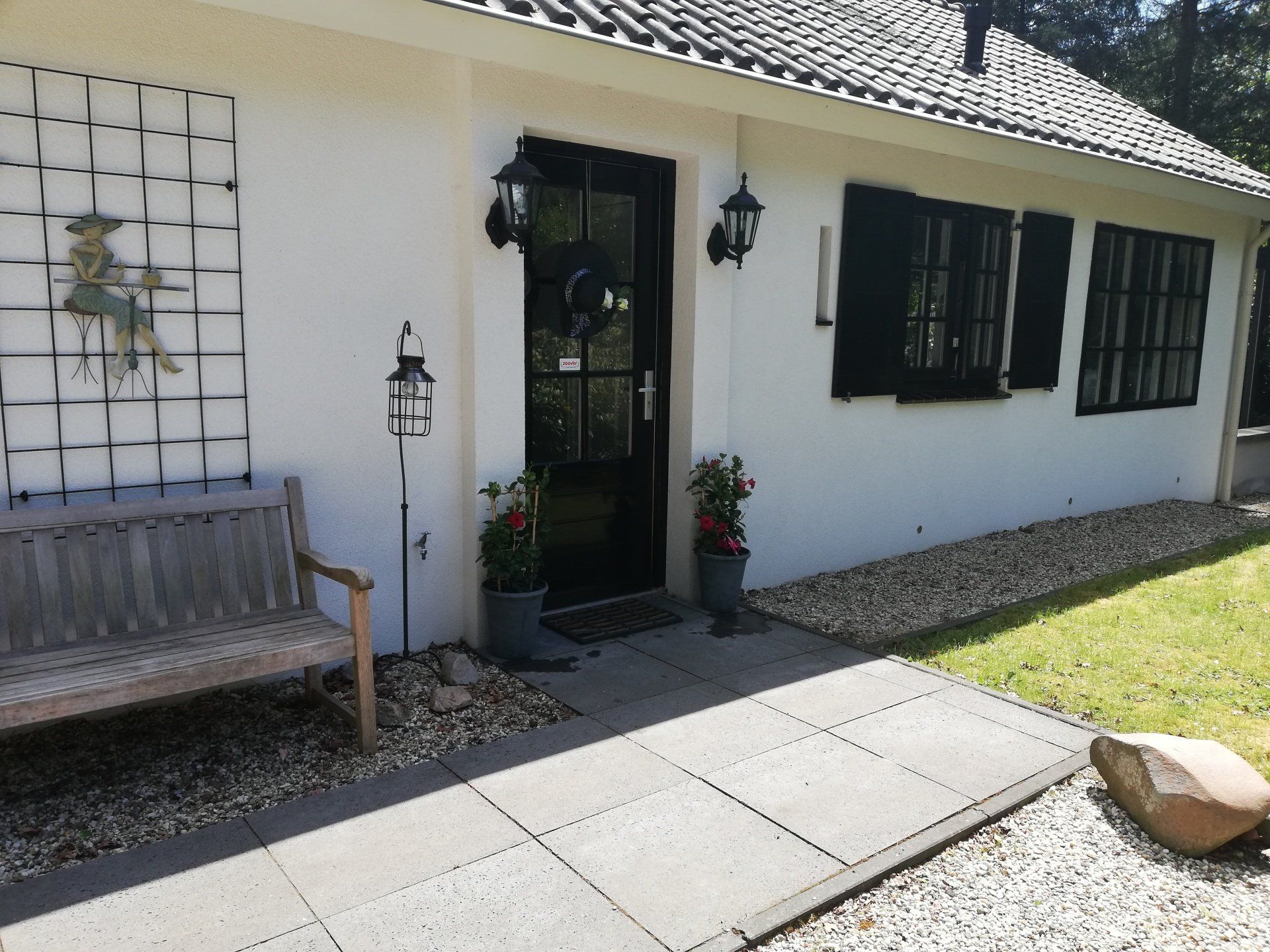 Wit huis met zwarte deur, luiken en lantaarn, plus een bankje en een stenen pad.
