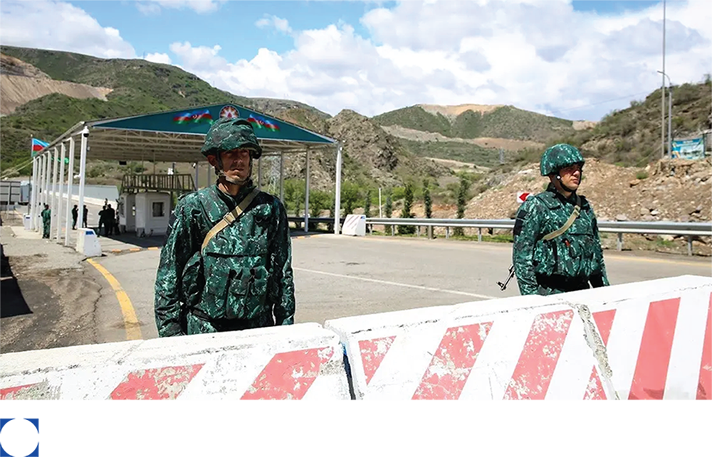 Two Azerbaijani soldiers in camouflage standing at a checkpoint with a mountainous landscape behind them.