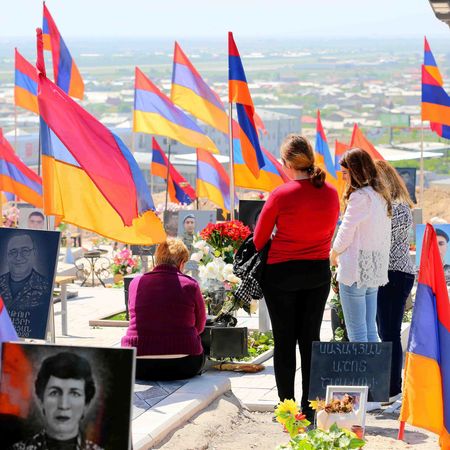 People at a gravesite with Armenian flags, looking at photos and flowers. Sunny day, outdoor setting.