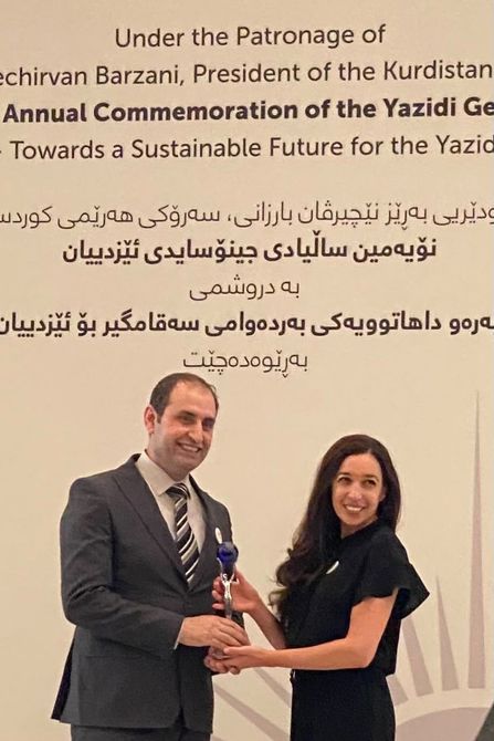 Haidar Elias  in suit presenting an award to a Lynn Zovighian. Background features commemorative text in English and Kurdish.