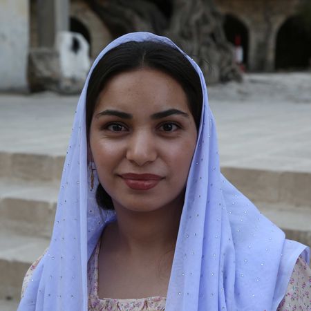 Woman in a light blue headscarf smiles in front of a stone building.
