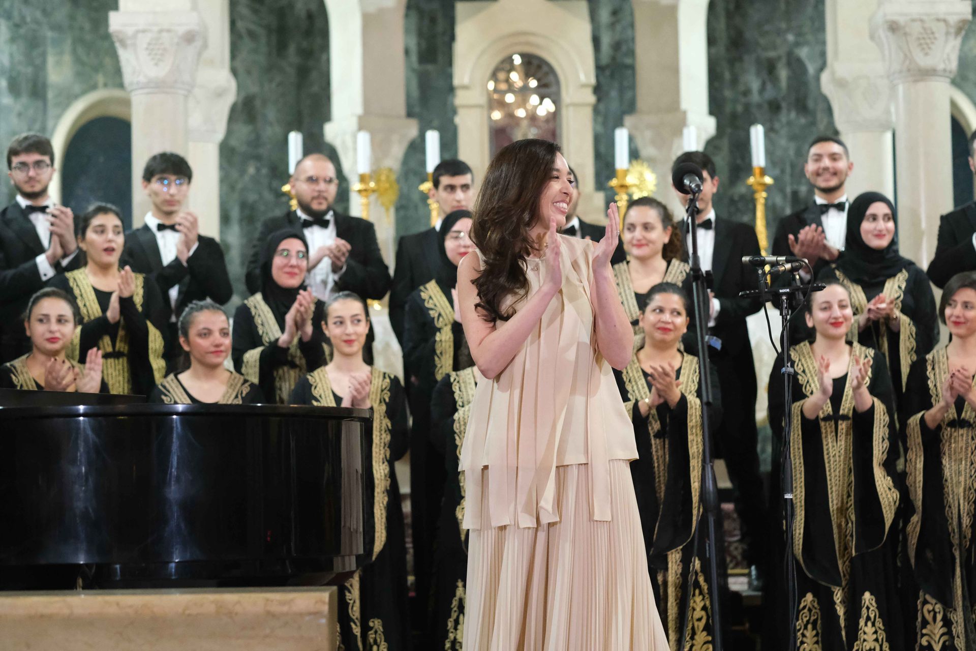 Lynn Zovighian in a beige dress clapping, welcomes the Fayha National Choir on stage at the concert “Giving Voice to Music.”
