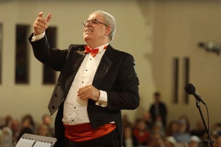 Maestro Barkev in a tuxedo and red bow tie with outstretched arms a conducting at Giving Voice to Music concert.
