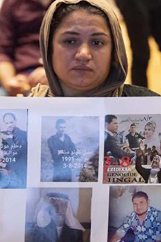 Woman wearing a veil holding photos, looking somber. Some photos show dates, other images, likely victims. Indoors setting.