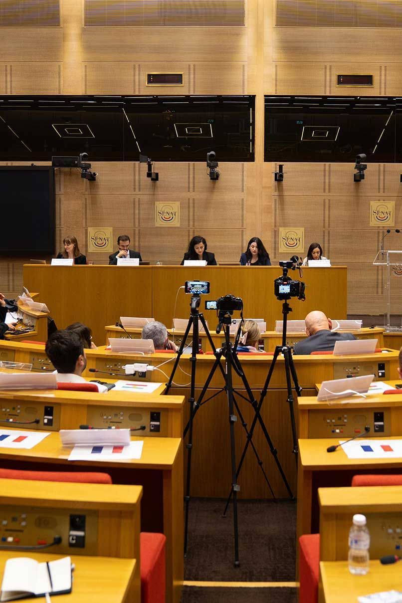 Conference room with people seated at a podium, cameras in the foreground, during a session at Palace of Luxembourg.