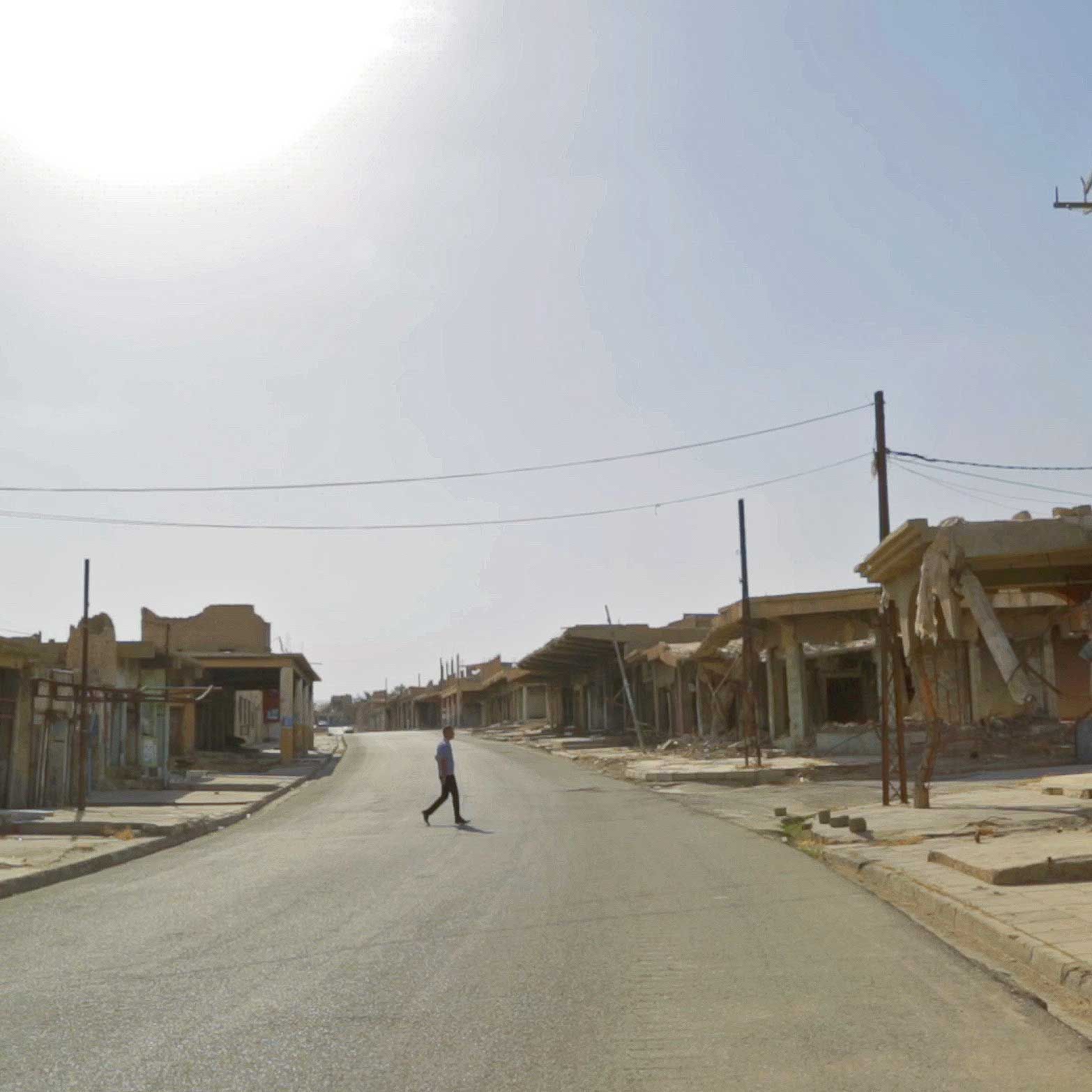 A person walks across a deserted road lined with damaged buildings under a bright sky.