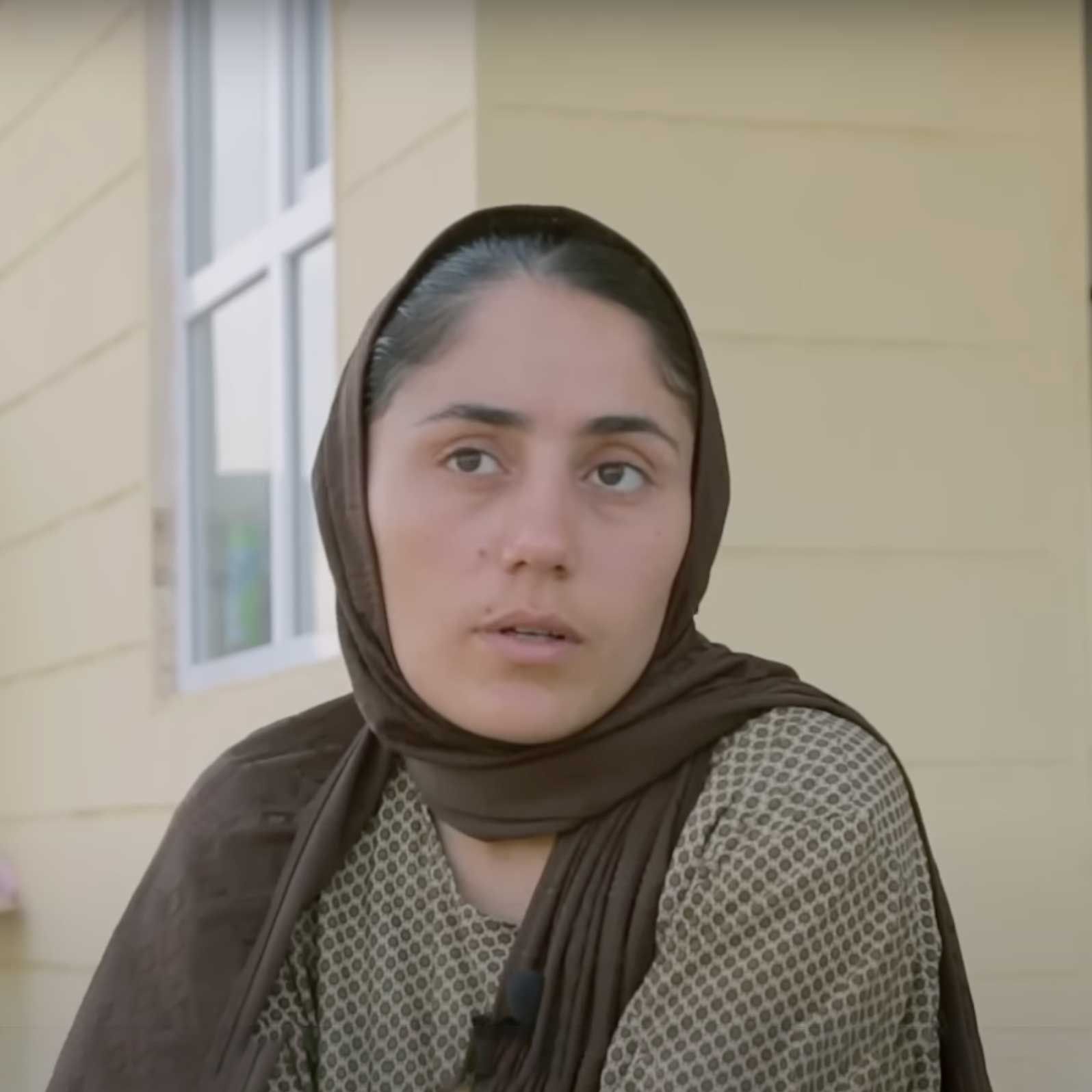 Woman in brown headscarf and patterned top, speaking outdoors in front of a window.