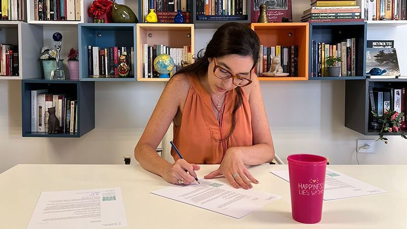 Lynn Zovighian wearing glasses writing at a desk, papers and pink cup. Shelves with books and objects in the background.