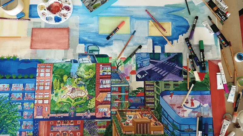Overhead view of an art table; watercolor painting of a Beirut city scene, brushes, markers, paint palette scattered.