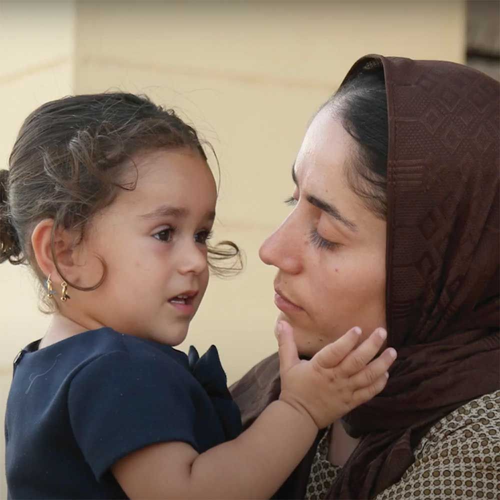 Woman holding a child, face-to-face. Both are looking toward each other, outdoors. Woman wears a brown headscarf.