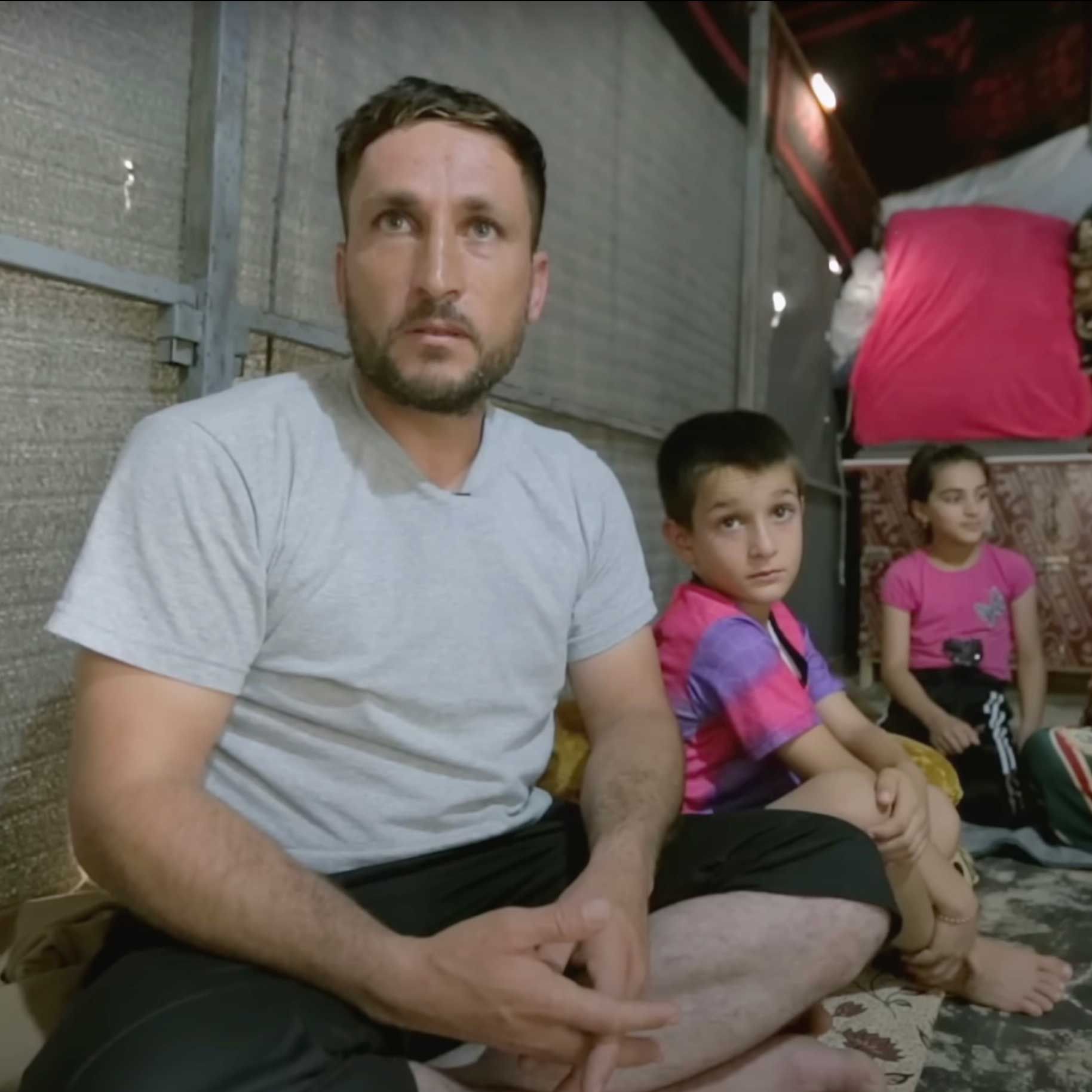 Man and two children sit indoors, looking forward. Man wears grey shirt, shorts. Boy in colorful shirt. Girl in pink.