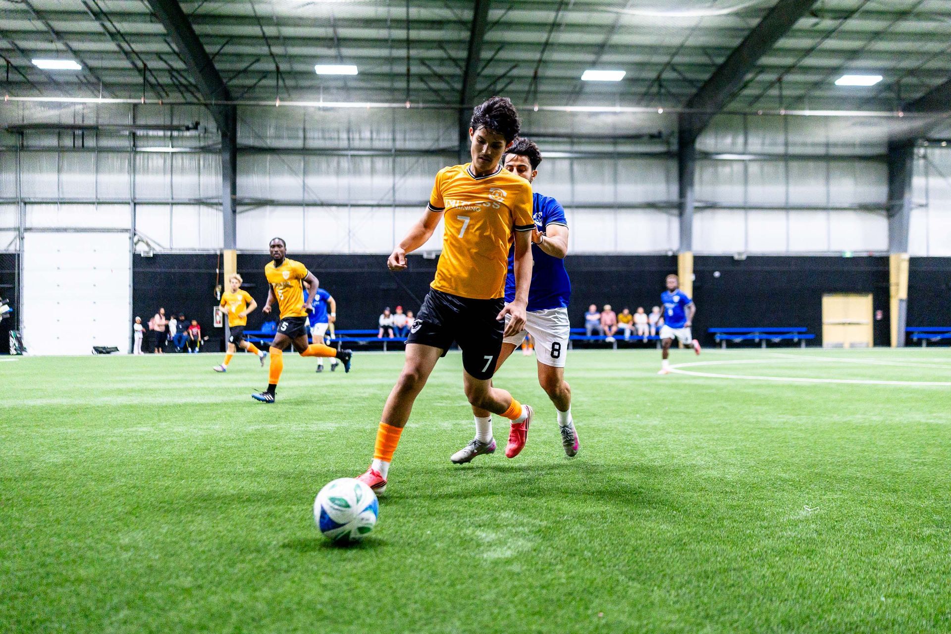 A player from Team Yellow and Team Blue chase a ball near the sidelines during Limitless Cup play in the warehouse like Viktoria Soccer Club indoor field.