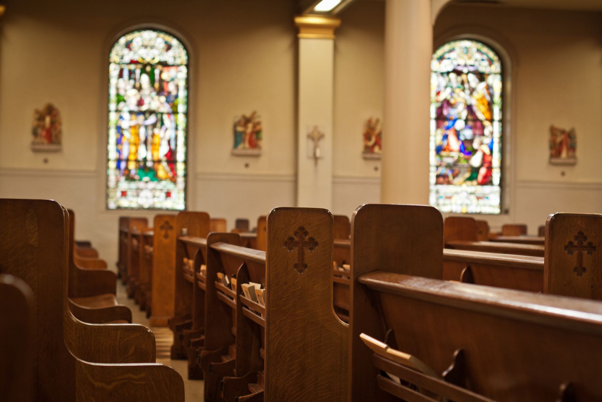 Rows of wooden pews in an empty church, with stained glass windows and a cross on the wall.