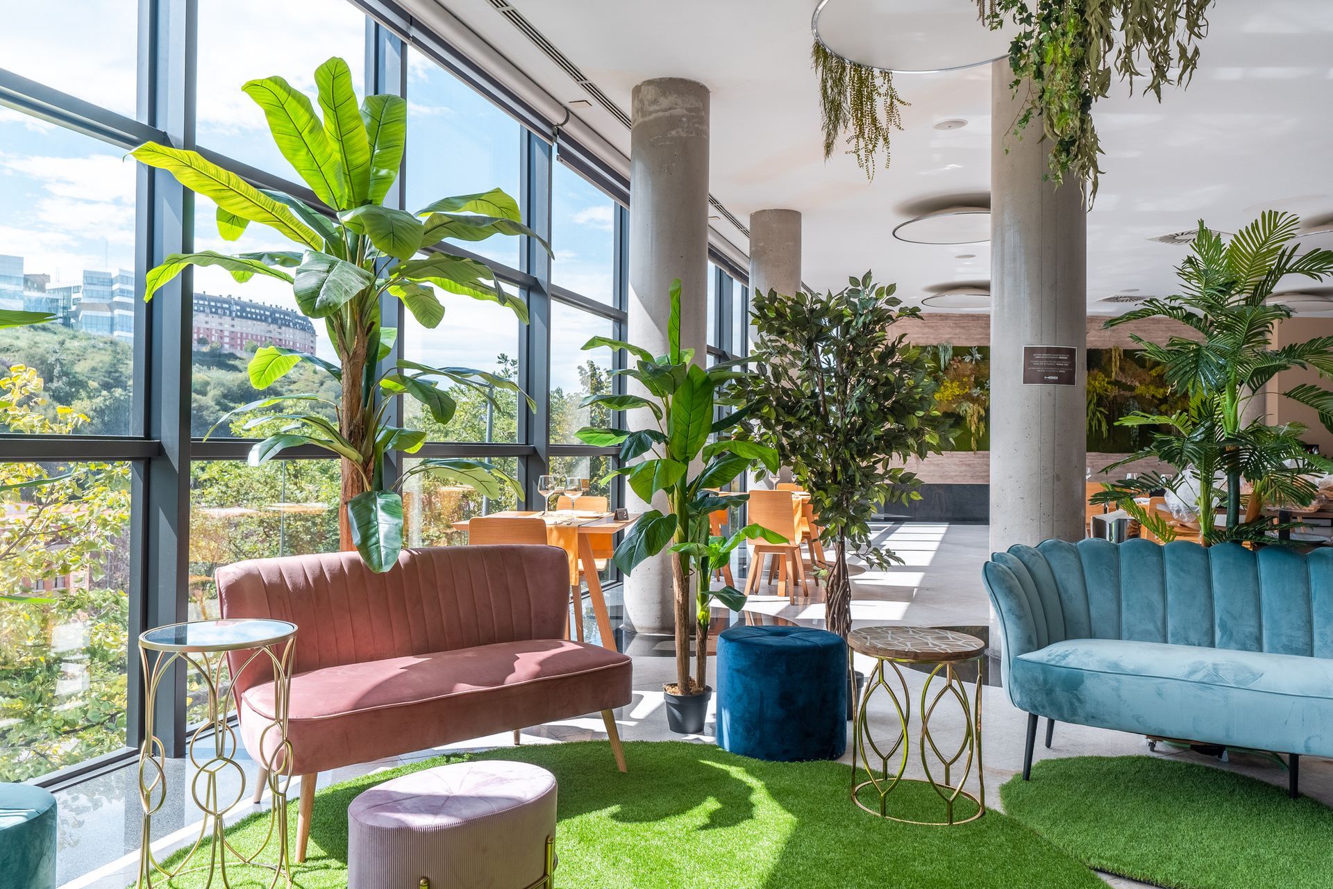 Lounge area with velvet sofas, plants, and large windows overlooking an outdoor view. Green turf rug.