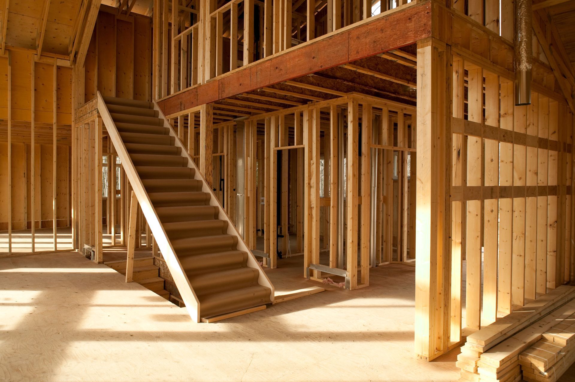 Interior of a house under construction; wooden frame walls, stairs, and flooring; bright sunlight.