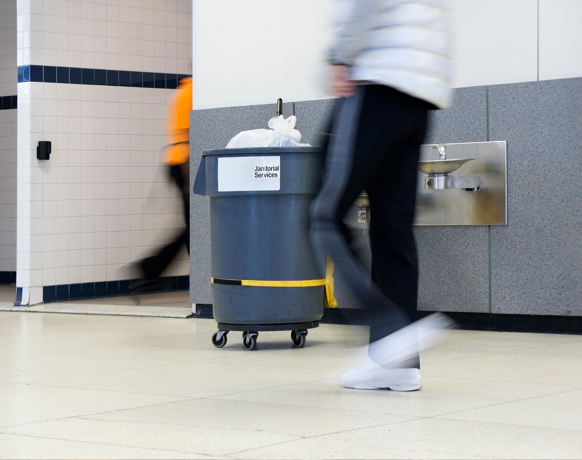People walking in a school hallway, blurred motion. Trash can on wheels, water fountain, and bathroom entrance visible.
