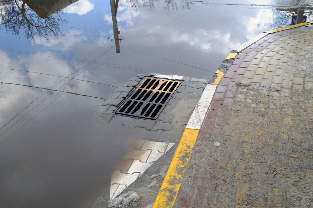 Una griglia di scolo parzialmente sommersa in una profonda pozzanghera su una strada asfaltata con un marciapiede dipinto di giallo e bianco.