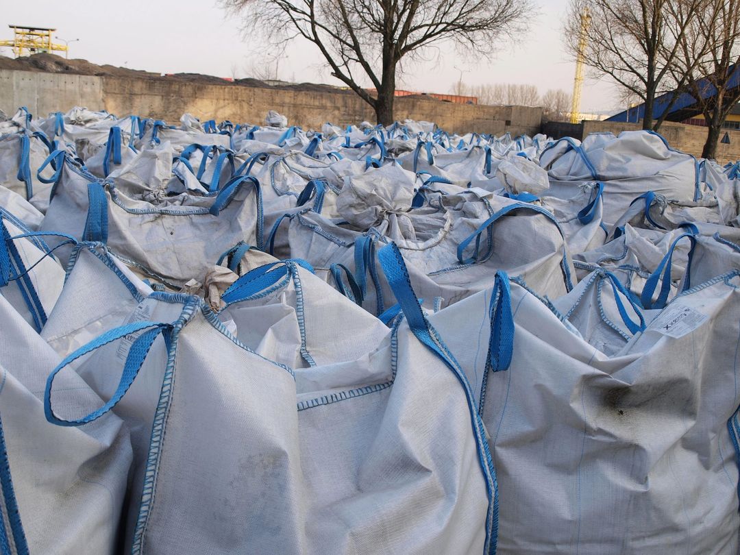 In un'area industriale all'aperto, numerose grandi borse portaoggetti bianche in tessuto con manici blu sono disposte in file.