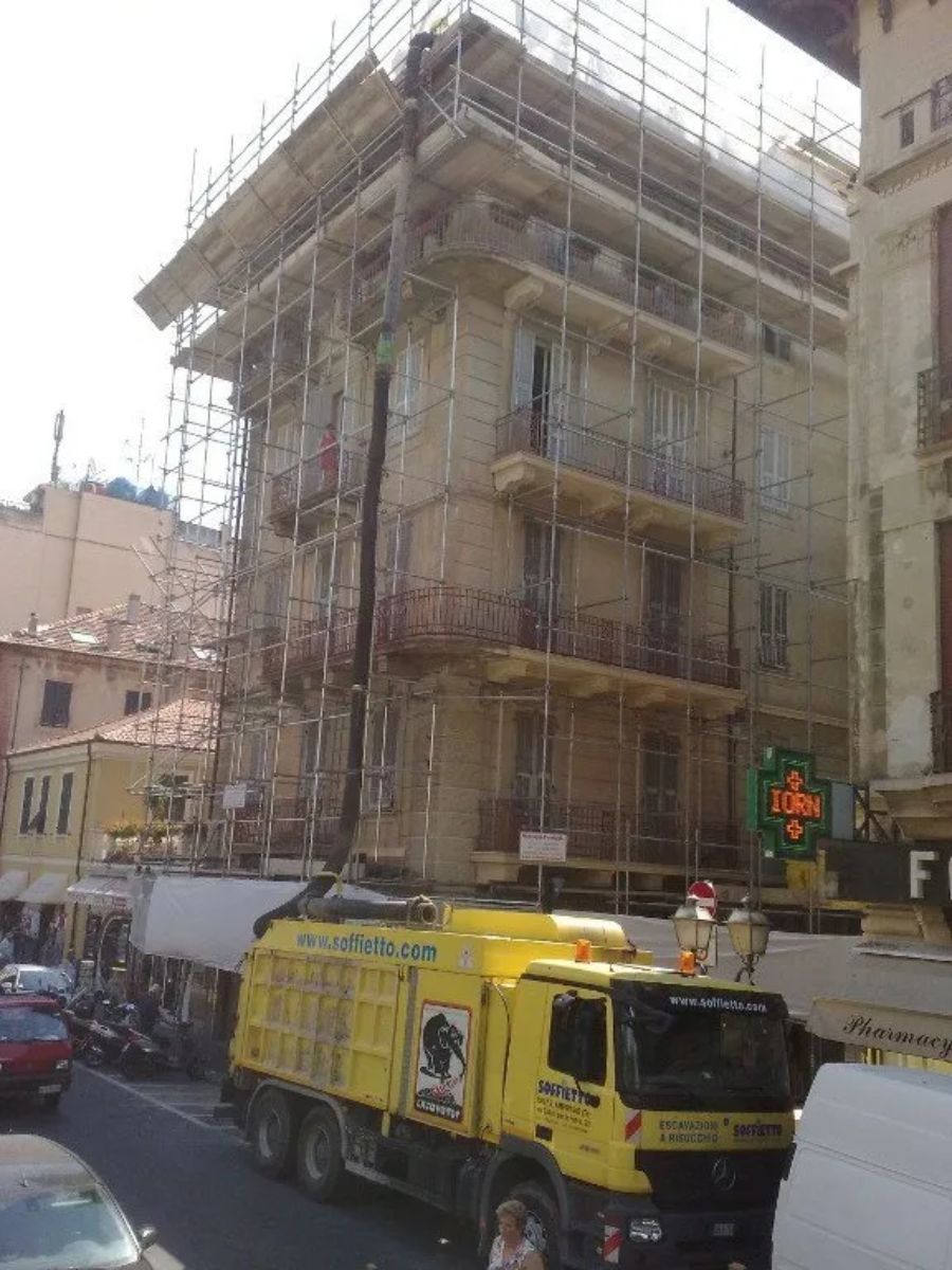 Un camion da cantiere giallo parcheggiato in una stretta via cittadina di fronte a un edificio a più piani ricoperto di impalcature.