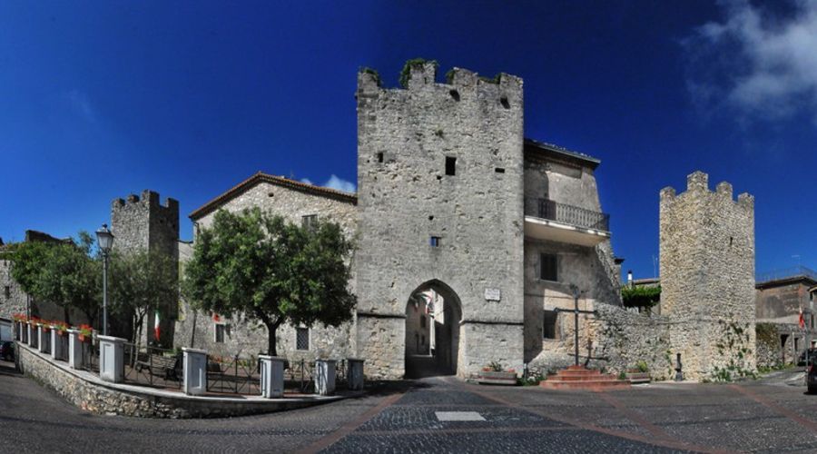 Una ripresa grandangolare di una storica fortezza in pietra con due torri, un portale ad arco e alberi sullo sfondo di un cielo azzurro brillante.