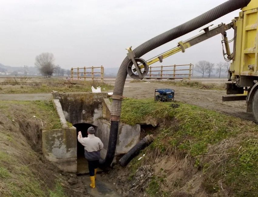 Un operaio con gli stivali gialli utilizza il tubo di un camion aspiratore per pulire un canale di scolo in cemento all'aperto in un campo erboso.