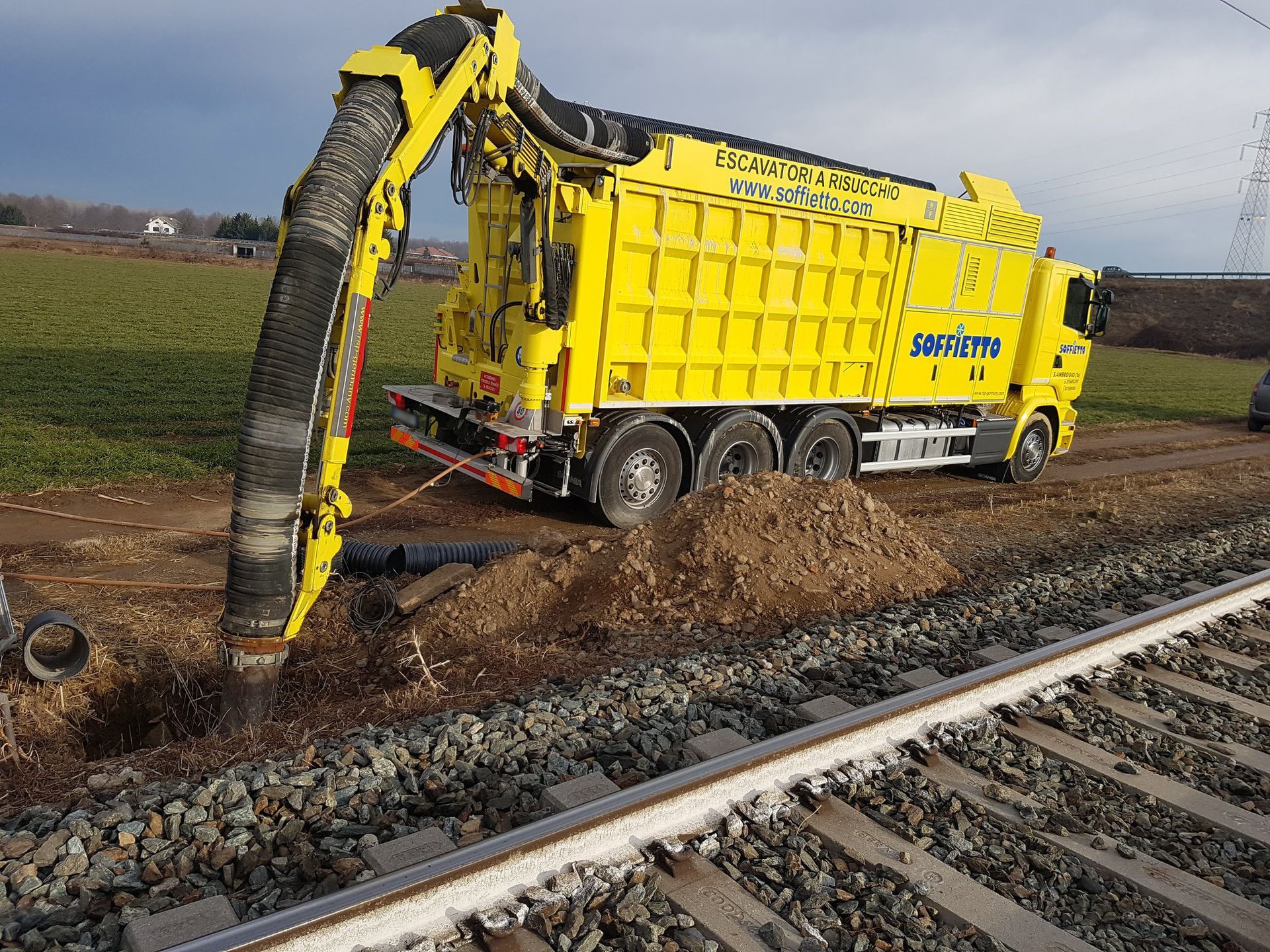 Un camion escavatore a risucchio di colore giallo, dotato di un grande tubo flessibile, pulisce il terreno lungo un binario ferroviario.