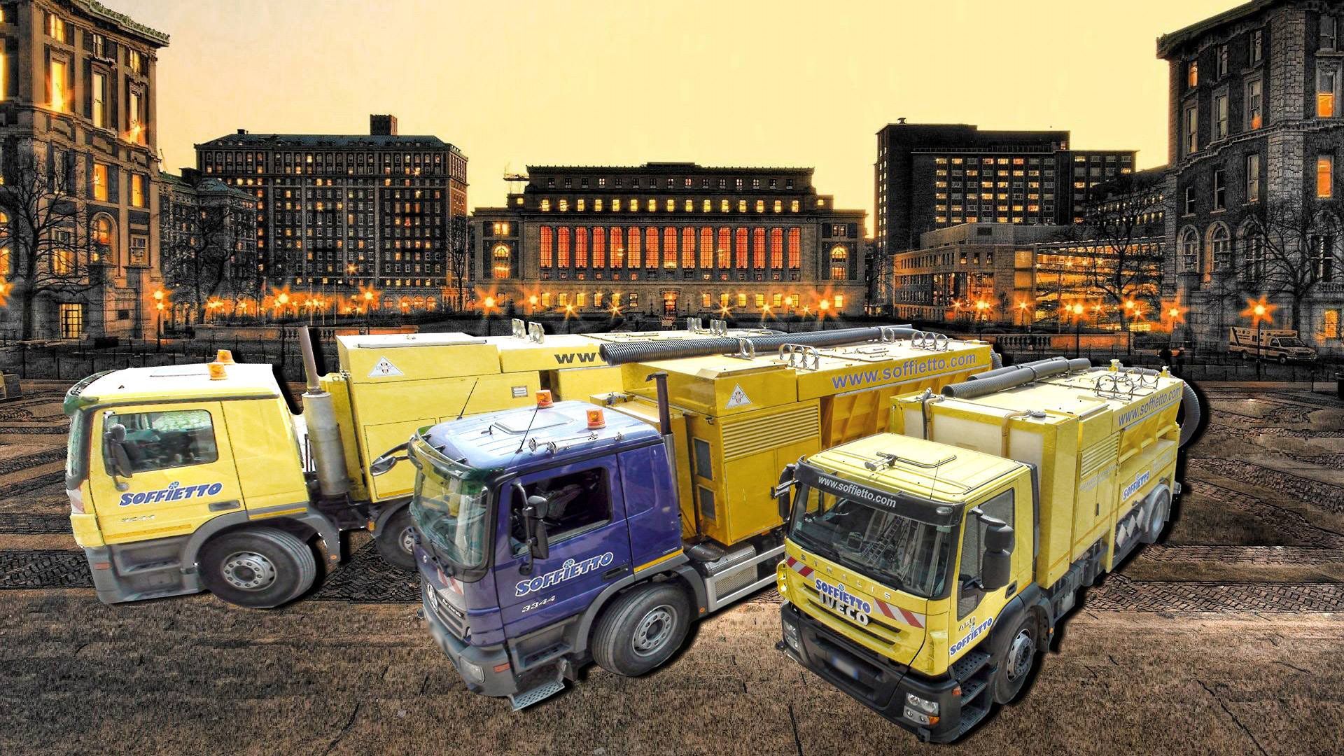 Tre camion da cantiere, uno viola e due gialli, parcheggiati su un terreno irregolare con uno sfondo di edifici cittadini.