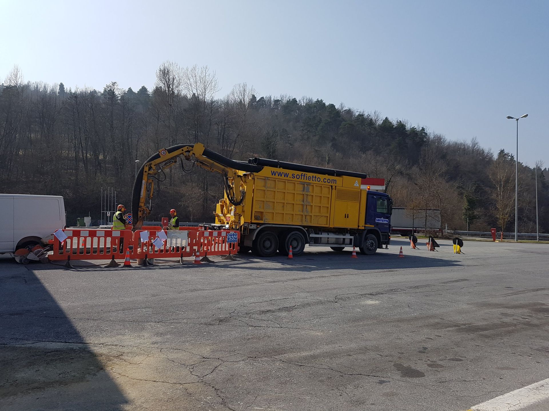 Un camion aspiratore giallo con un tubo flessibile estensibile è parcheggiato in un piazzale asfaltato dietro barriere di sicurezza in plastica arancione.