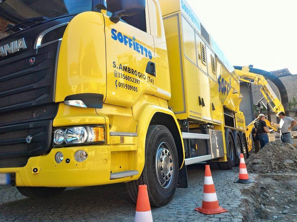 Un camion escavatore a risucchio Scania di colore giallo brillante è parcheggiato su una strada acciottolata, con operai e attrezzature da costruzione a bordo.