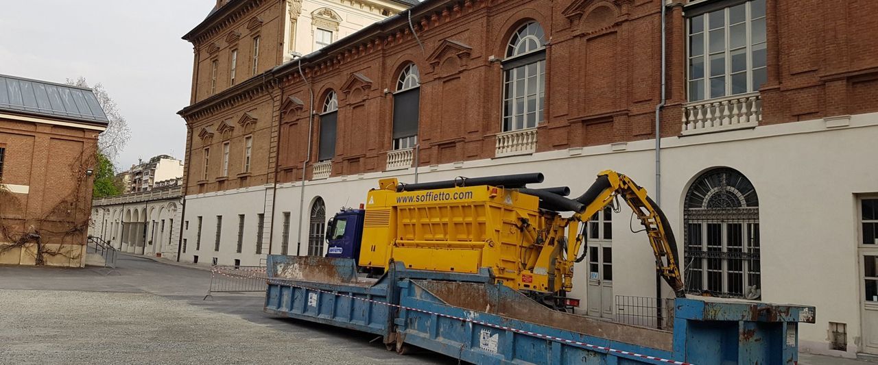 Un veicolo da cantiere giallo con un braccio robotico è parcheggiato vicino a un cassonetto blu all'esterno di un edificio storico in mattoni e bianco.