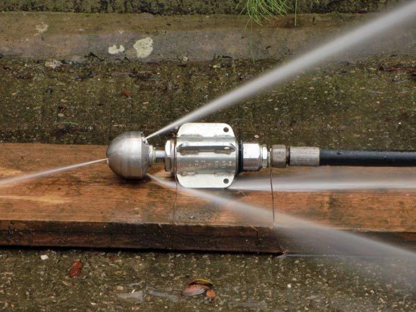 Un ugello metallico per la pulizia delle fognature spruzza getti d'acqua ad alta pressione in diverse direzioni, mentre è appoggiato su una tavola di legno.