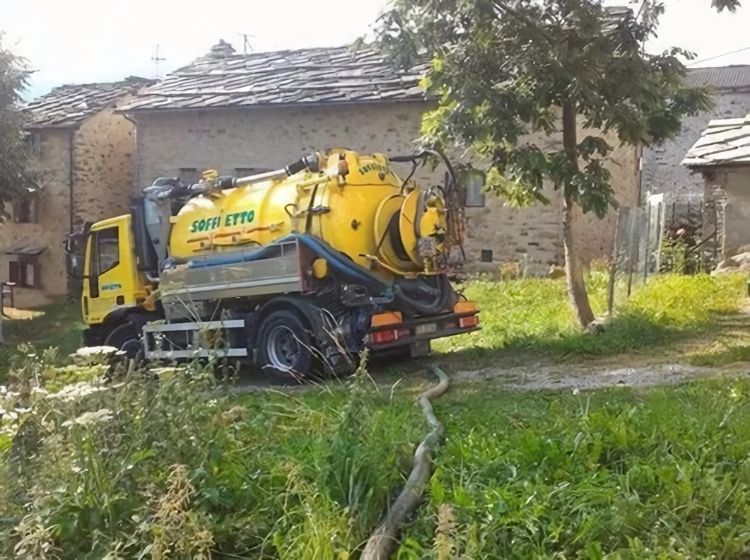 Un camion cisterna industriale giallo brillante per lo svuotamento di fosse settiche è parcheggiato su un'area erbosa di fronte ad edifici in pietra.