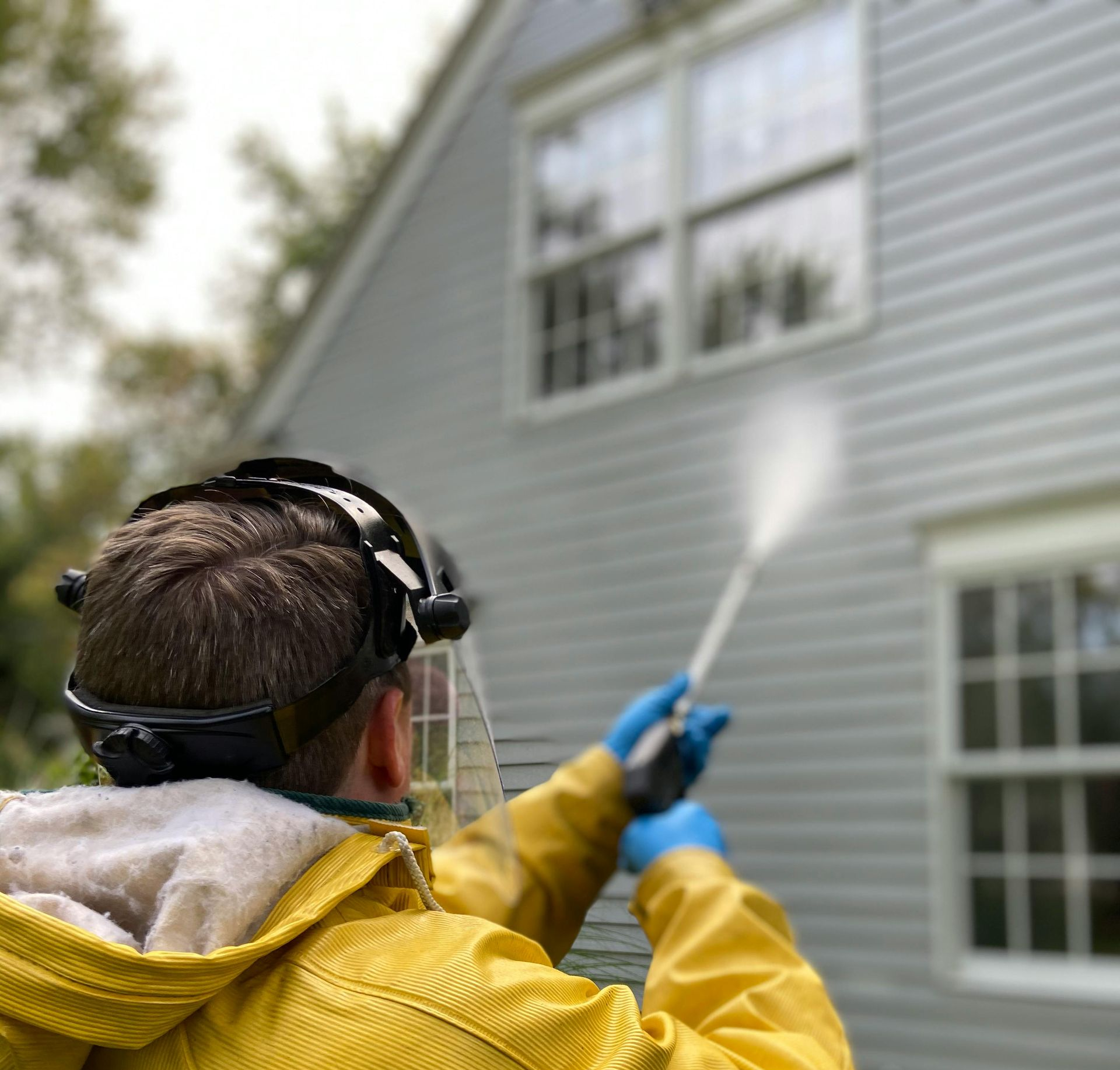Person in yellow raincoat pressure washing a house exterior near windows
