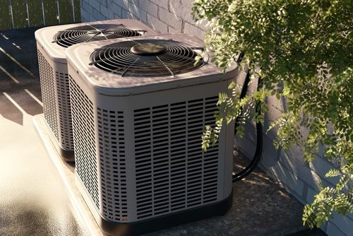 Two outdoor air conditioner units beside a house wall in sunlight, partly shaded by tree branches