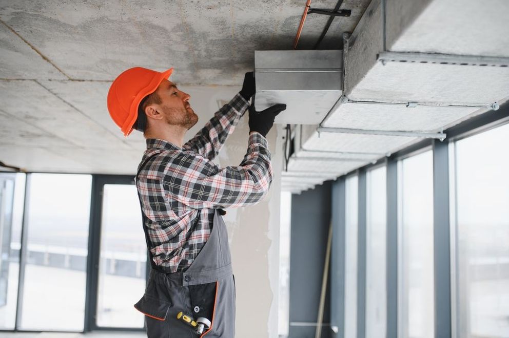 Construction worker installing ducting on a ceiling in an unfinished building, wearing an orange hard hat.
