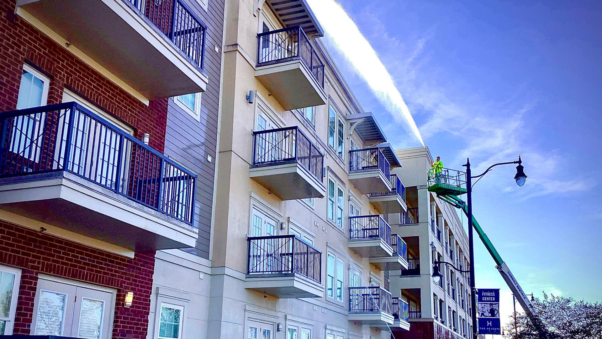 Apartment building with balconies; worker in lift, painting side of building; bright blue sky.