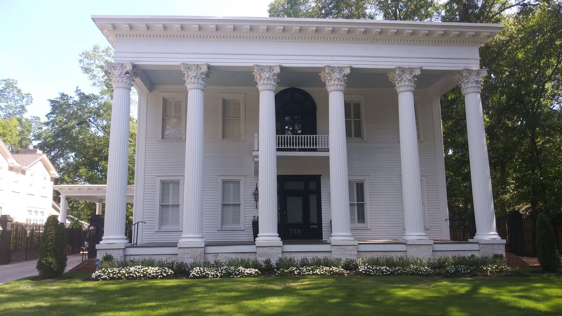 A two-story white neoclassical mansion with six tall Corinthian columns, a central balcony, and a green front lawn.