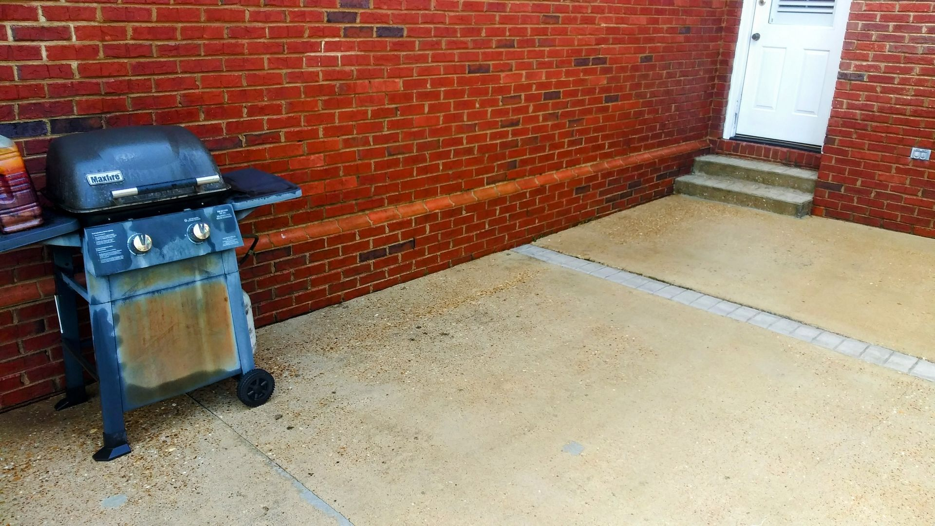Barbecue grill on a concrete patio next to a red brick wall and steps leading to a white door.